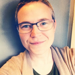 Photo of short-haired Alex wearing glasses and a straw-colored cardigan.