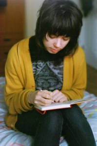 Photo of a dark-haired zinester wearing a yellow cardigan and writing in a notebook.