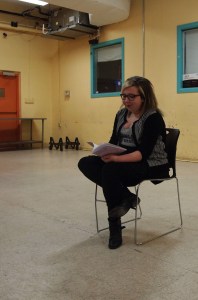 JC seated in a plastic chair reading from their zine.