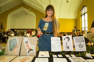 Maggie Negrete, a long blond-haired person, standing behind a row of their zines at a zine fest.