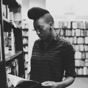 B&w photo of Sy looking down at a book amidst book-filled shelves.
